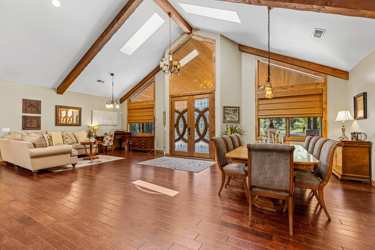 An open-plan dining and living area filled with natural light from large windows. It features a substantial dining table, a comfortable sofa, and rustic wooden beams overhead—fostering a welcoming space for dining, socializing, and relaxing.