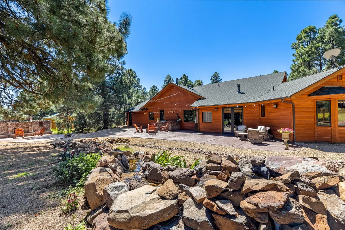A tranquil exterior scene featuring a wooden house with patio seating, a landscaped stone border, and mature trees in the background. The patio offers a restful space for enjoying the outdoors, while the natural surroundings create a peaceful atmosphere ideal for relaxation and nature appreciation.