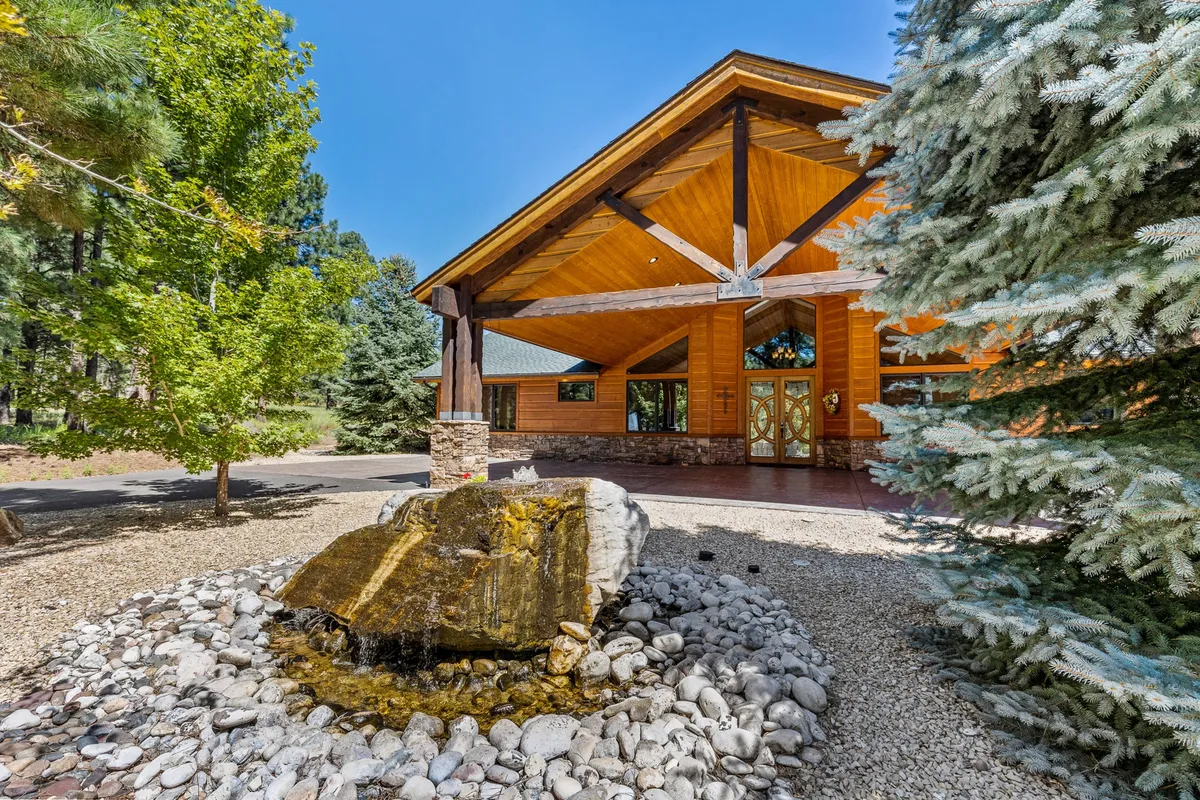 The image shows a spacious exterior with a large wooden entrance, a tranquil water feature, stone pathways, and lush greenery surrounding the entry, suggesting both privacy and a serene outdoor escape. The setting evokes feelings of arriving at a secluded, welcoming getaway, perfect for relaxing evenings or romantic walks.