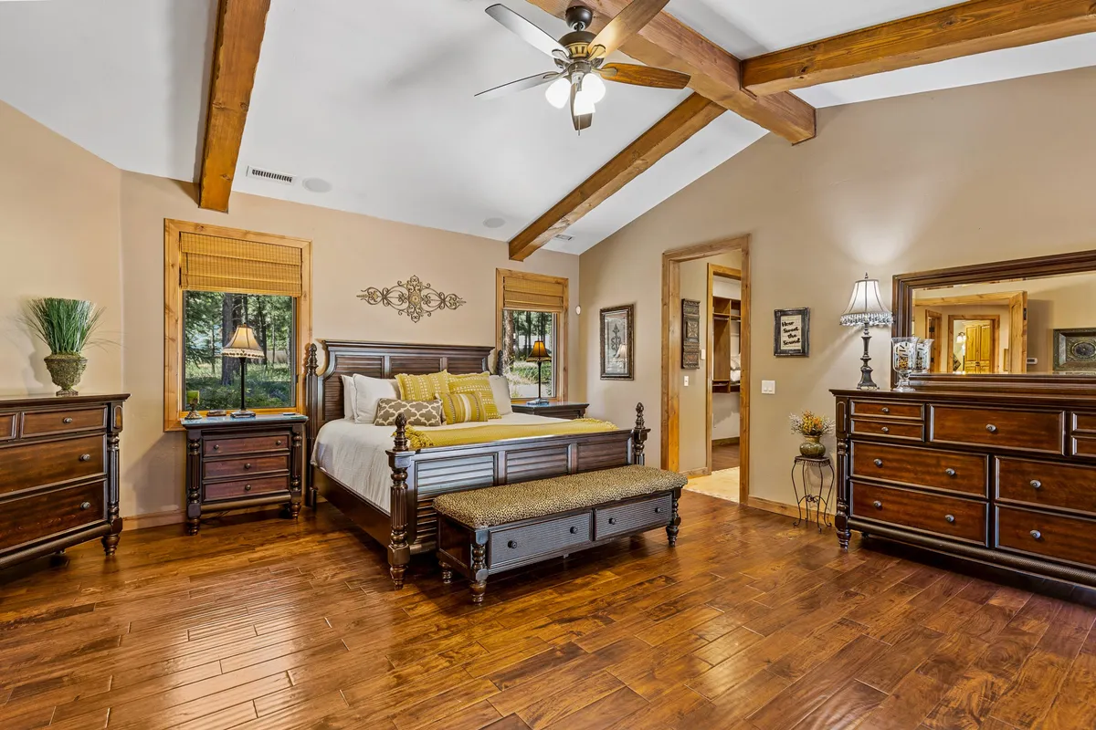 A tranquil guest bedroom featuring a queen-size bed, a dresser, a mirror, and a large window letting in morning sunlight. The space is decorated with calming artwork and exudes a restful, homey ambiance.