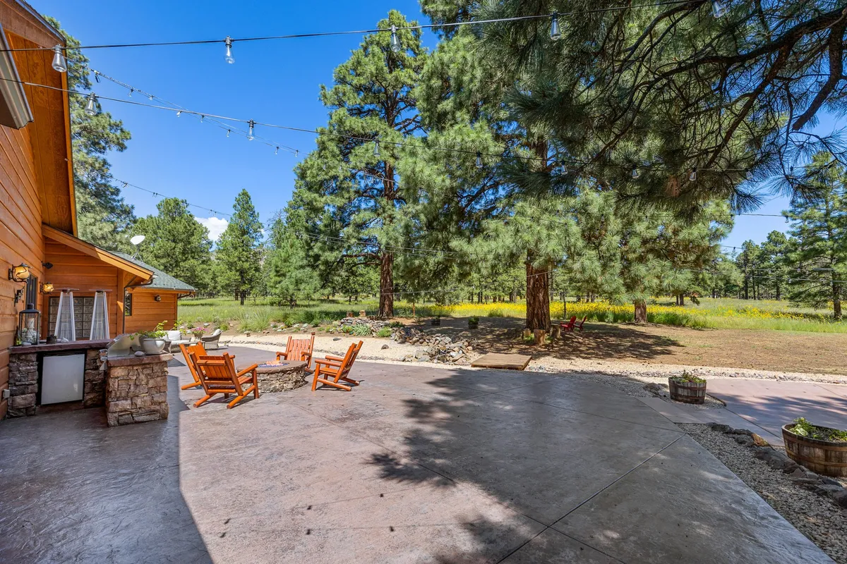 A tranquil patio area is set with wooden chairs and surrounded by green trees, illuminated by decorative lights. The space feels open to the sky, ideal for stargazing, morning coffee, or relaxing after a day of adventures. The lush setting hints at the peaceful transition from home to nature.