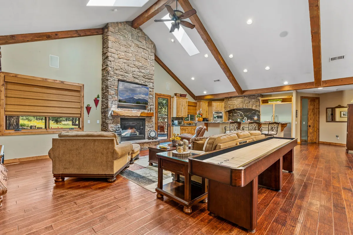 A warm and inviting living area featuring a stone fireplace, large windows ushering in natural light, exposed wooden beams, a comfortable seating area, and a shuffleboard table. The scene captures the essence of family gatherings, relaxed evenings by the fire, and lively game nights.