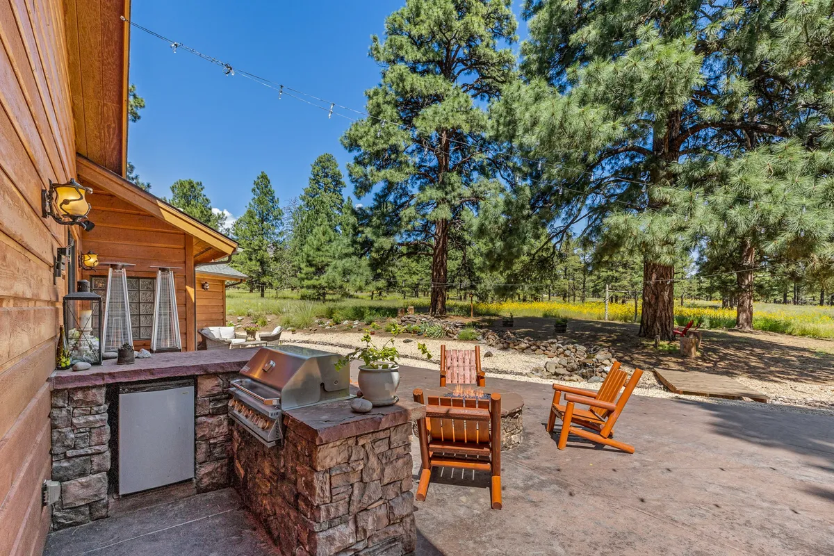 A wooden deck features an outdoor grill and comfortable wooden chairs under the canopy of tall pine trees. The area exudes a casual ambiance, ideal for barbecues and social gatherings, with string lights providing a festive touch.