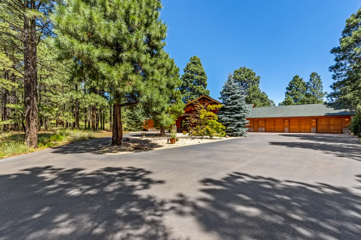 A rustic log cabin sits nestled among tall trees, with a driveway leading up to the home and thick forest on all sides. The porch offers a direct connection to the outdoors, signaling the immediate proximity to natural adventures. The surrounding trees emphasize the home's integration with the wild landscape of Northern Arizona.