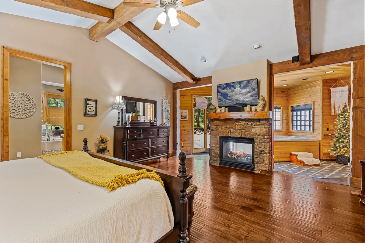 A serene master suite featuring a plush bed, a glowing stone fireplace, wooden beams overhead, a dressing mirror, and large windows that fill the space with natural light.