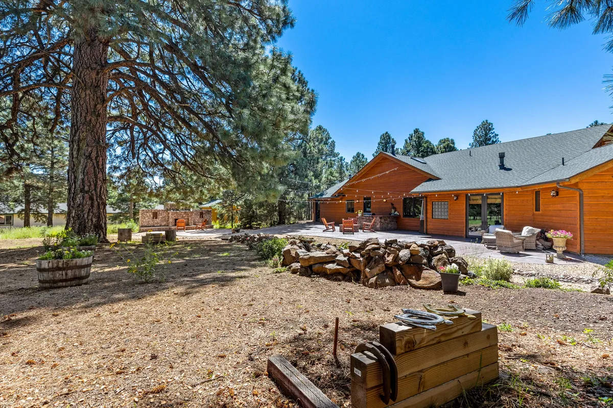 This exterior image shows a cozy stone fire pit with an outdoor seating area set beside a wooden house and a large pine tree. The area is perfect for evening gatherings, s’mores, or stargazing, surrounded by nature.
