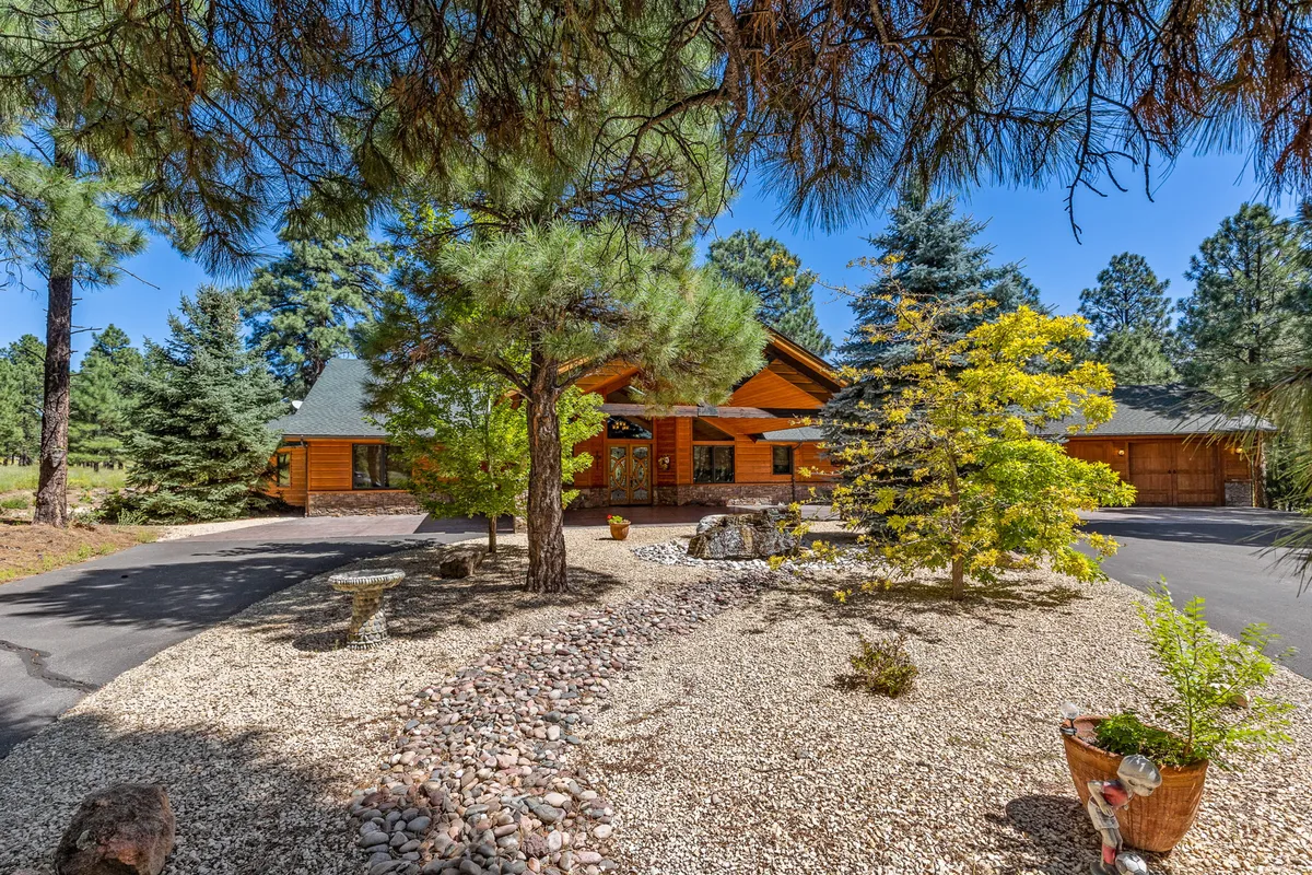 The log cabin facade is framed by a stone pathway, colorful shrubs, and a welcoming front door with decorative touches. The lush landscaping and cabin architecture evoke a sense of mountain tranquility and connection to the outdoors.