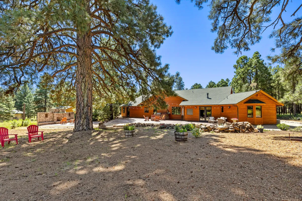 A rustic patio set with red outdoor chairs encircles a stone fire pit beneath a towering pine. The wooden house and forested landscape beyond frame the setting, offering a tranquil space for storytelling, barbecues, and winter activities.