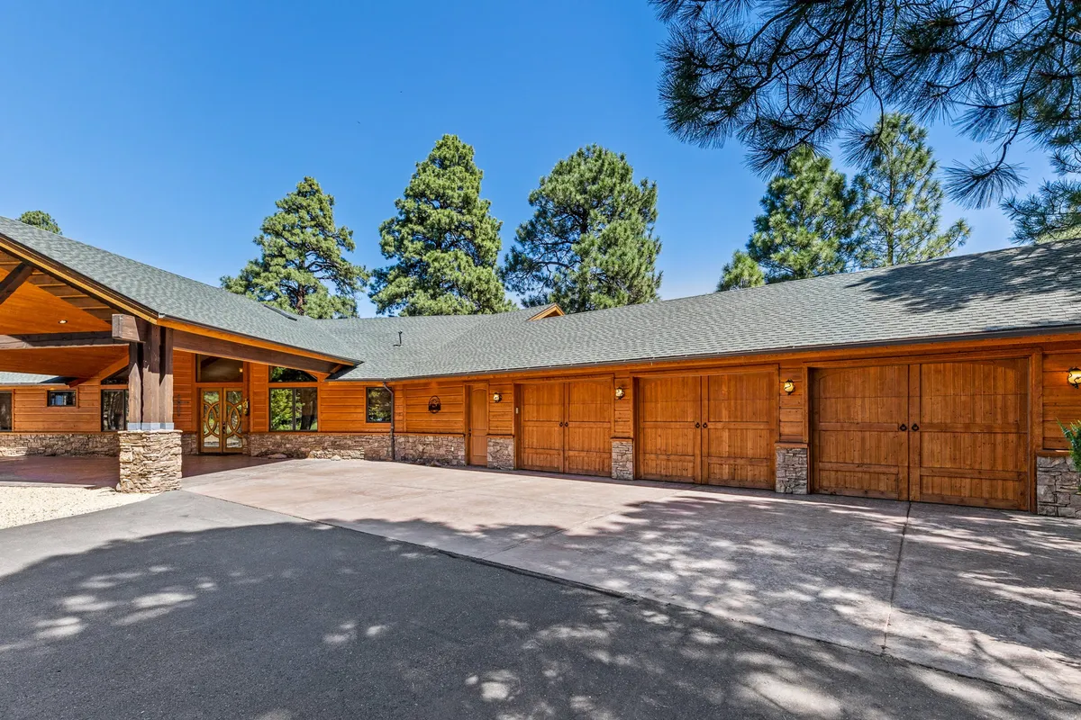 This exterior scene captures the main entrance with wooden garage doors, a stone façade, and towering pine trees encircling the property. The setting offers a sense of seclusion and connection to nature just minutes from the city.